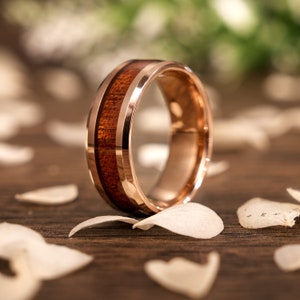 May include: A rose gold tungsten carbide ring with a brown wood inlay. The ring is on a wooden surface with white flower petals.