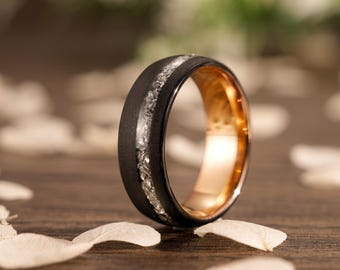 May include: A black ring with a gold interior and a band of clear, sparkling stones. The ring is set against a blurred background with white petals, creating a contrast of textures and colors. The ring's design is modern and elegant.
