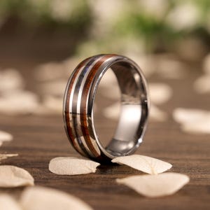 May include: A silver tungsten carbide ring with a brown wood inlay and a silver and blue abalone shell inlay. The ring is on a wooden surface with white flower petals.