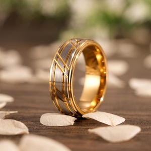 Könnte beinhalten: Ein gold- und silberfarbener Ring mit geometrischem Design. Der Ring hat ein goldfarbenes Innen- und Außendesign mit einem silberfarbenen Band in der Mitte. Das silberfarbene Band hat ein Kreuzmuster. Der Ring liegt auf einer Holzoberfläche.