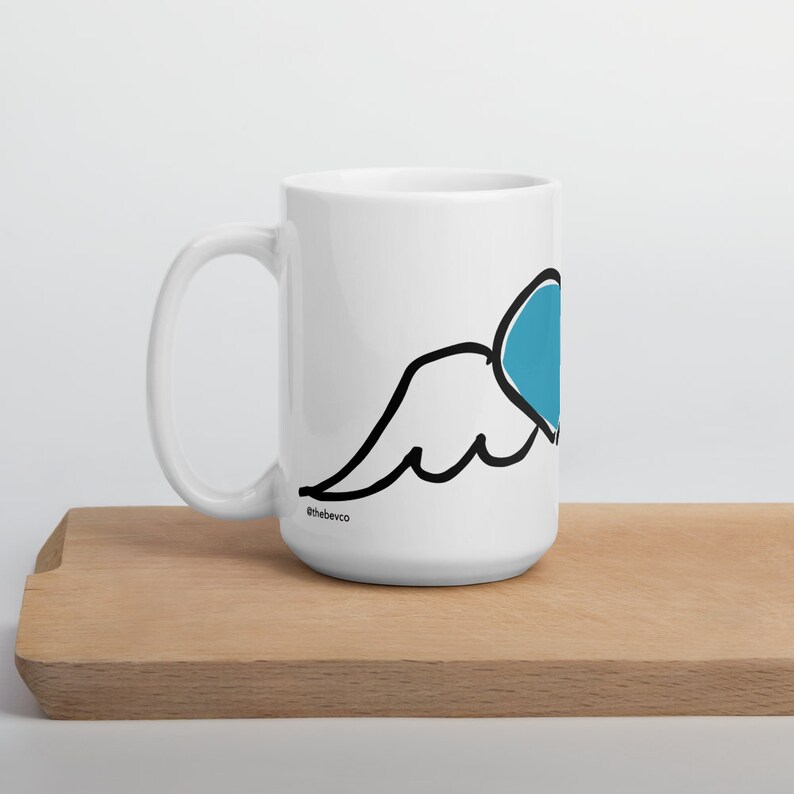 May include: White ceramic mug with a black outline of a wing and a blue heart. The mug has the text "@thebevco" on the bottom.
