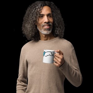 May include: A man with long, gray hair is holding a white mug with a blue heart and a black mustache design.