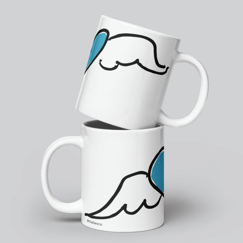 May include: Two white ceramic mugs stacked on top of each other. The mug on top has a blue heart with wings drawn on it. The mug on the bottom has the same design but the heart is on the right side of the mug. The text "@thebevco" is printed on the bottom mug.