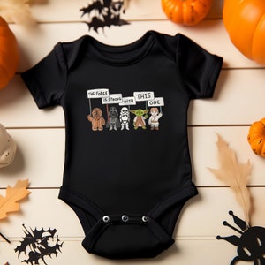 May include: Black baby onesie with a Star Wars graphic. The graphic features Chewbacca, Darth Vader, a Stormtrooper, Yoda, and Luke Skywalker holding signs that say "The Force is Strong with This One".