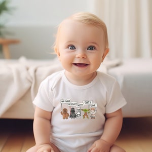 May include: A white baby onesie with a graphic of Star Wars characters holding signs that say "The Force is strong with this one".
