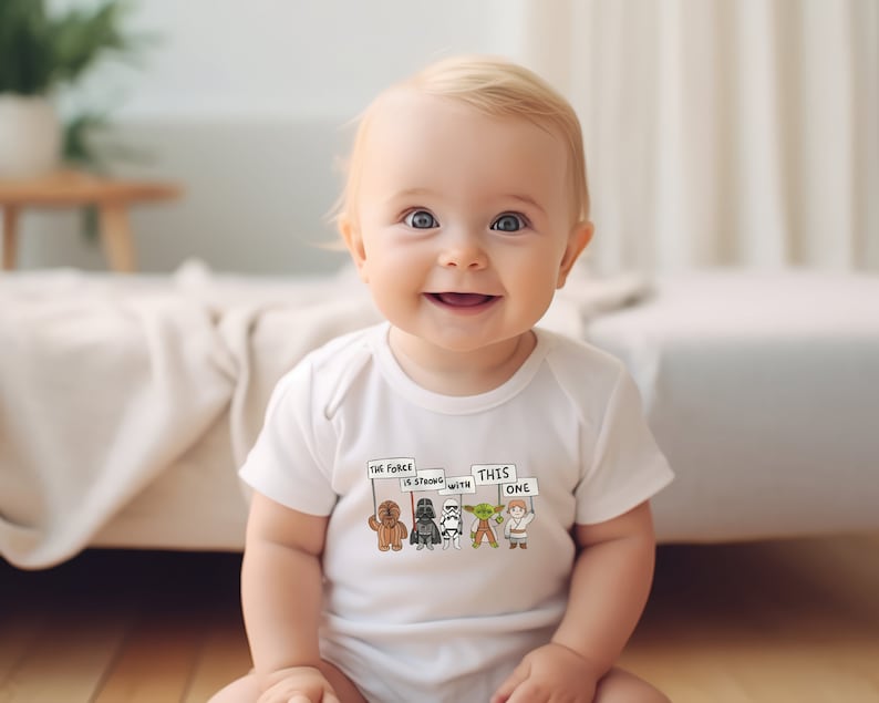 May include: A white baby onesie with a graphic of Star Wars characters holding signs that say "The Force is strong with this one".