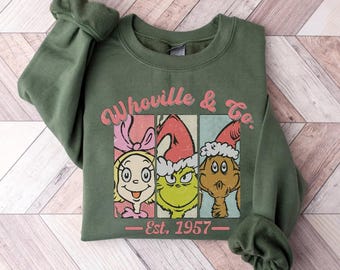 May include: Military green sweatshirt featuring a graphic of characters from Whoville, with the text "Whoville & Co." and "Est. 1957" in a retro style. The design includes three cartoon faces in separate panels, each wearing a Santa hat.
