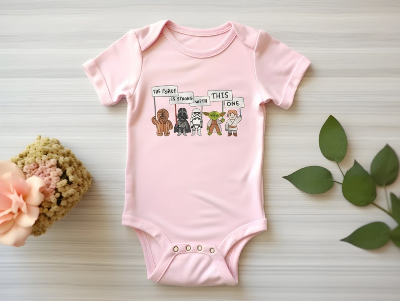 May include: Pink baby onesie with a graphic of Star Wars characters holding signs that say "The Force is Strong with This One".