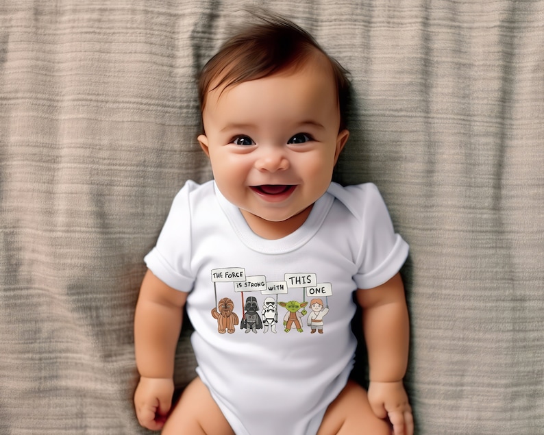 May include: A white baby onesie with a graphic of Star Wars characters holding signs that say "The Force is Strong with This One".