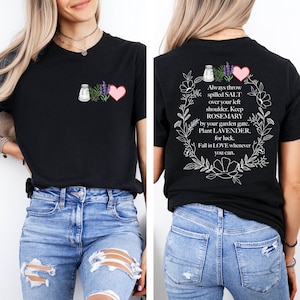 May include: Black t-shirt with a graphic of a salt shaker, lavender, and a pink heart on the front. The back of the shirt features a floral wreath with text that reads, "Always throw spilled SALT over your left shoulder. Keep ROSEMARY by your garden gate. Plant LAVENDER, for luck. Fall in LOVE whenever you can."