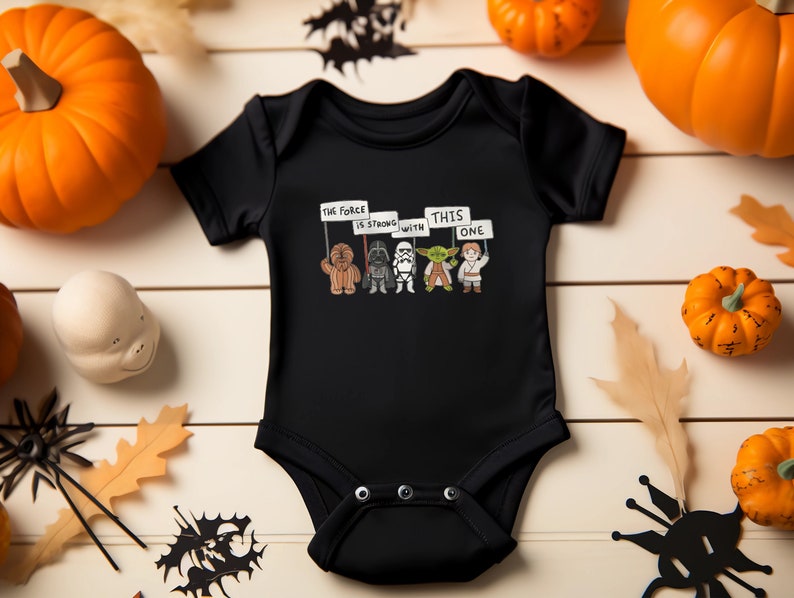 May include: Black baby onesie with a Star Wars graphic. The graphic features Chewbacca, Darth Vader, a Stormtrooper, Yoda, and Luke Skywalker holding signs that say "The Force is Strong with This One".