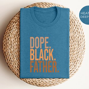 May include: A heather deep teal t-shirt with the words "DOPE. BLACK. FATHER." in orange lettering. The shirt is folded and placed on a woven, circular mat.