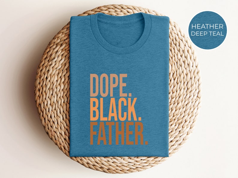 May include: A heather deep teal t-shirt with the words "DOPE. BLACK. FATHER." in orange lettering. The shirt is folded and placed on a woven, circular mat.
