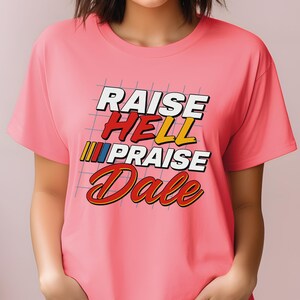 Pu&ograve; includere: T-shirt rosa chiaro con la scritta "RAISE HELL PRAISE Dale" in stile retr&ograve;. Il testo &egrave; in bianco, rosso, giallo e blu, su sfondo a griglia. La maglietta ha maniche corte.