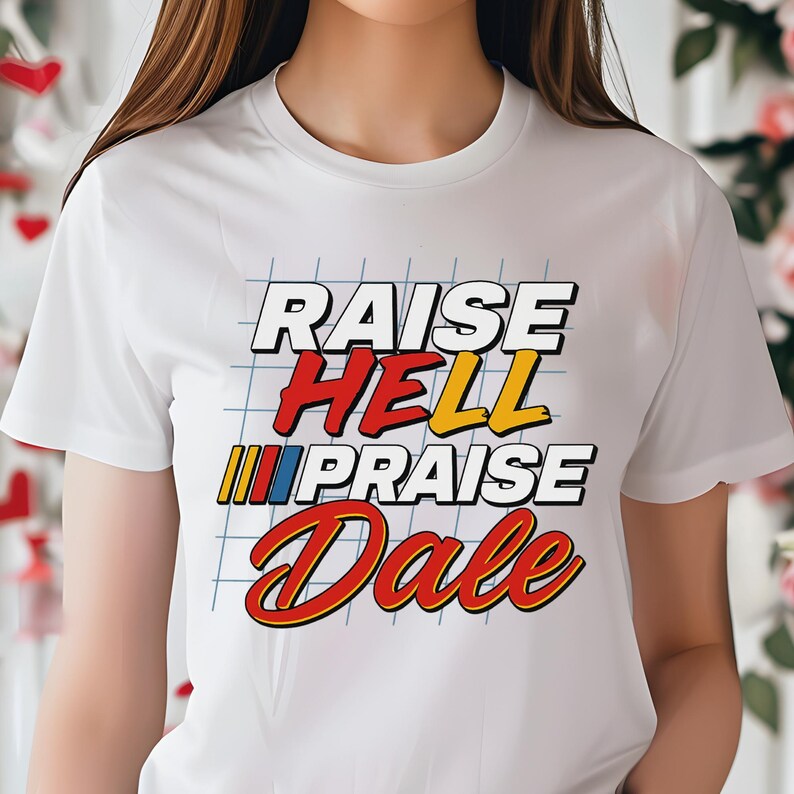 Pu&ograve; includere: T-shirt bianco con la scritta "RAISE HELL PRAISE Dale" in un font retr&ograve;. Il testo &egrave; in bianco, rosso, giallo e blu, con uno sfondo a griglia. Una maglietta grafica casual.