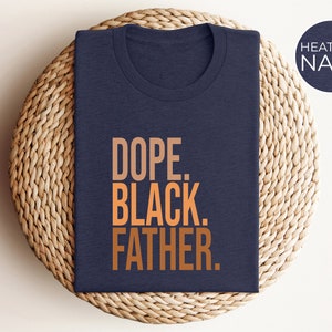 May include: Heather navy blue t-shirt with the words "DOPE. BLACK. FATHER." in a gradient orange font. The shirt is folded and placed on a woven, circular placemat. A navy blue circle with the words "HEATHER NAVY" is in the upper right corner.