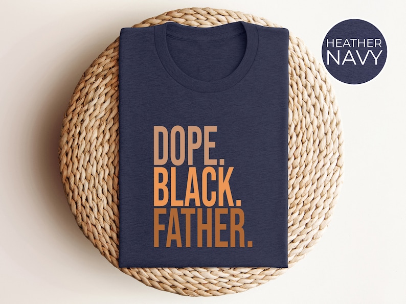 May include: Heather navy blue t-shirt with the words "DOPE. BLACK. FATHER." in a gradient orange font. The shirt is folded and placed on a woven, circular placemat. A navy blue circle with the words "HEATHER NAVY" is in the upper right corner.