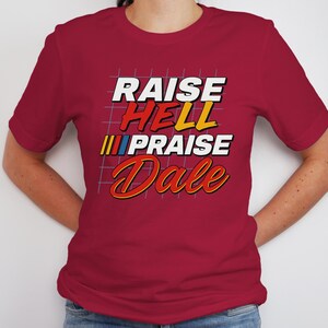 Pu&ograve; includere: Una maglietta bordeaux con la scritta "RAISE HELL PRAISE Dale" in un font retr&ograve;. Il testo &egrave; in bianco, rosso, giallo e blu. La maglietta ha maniche corte e girocollo.