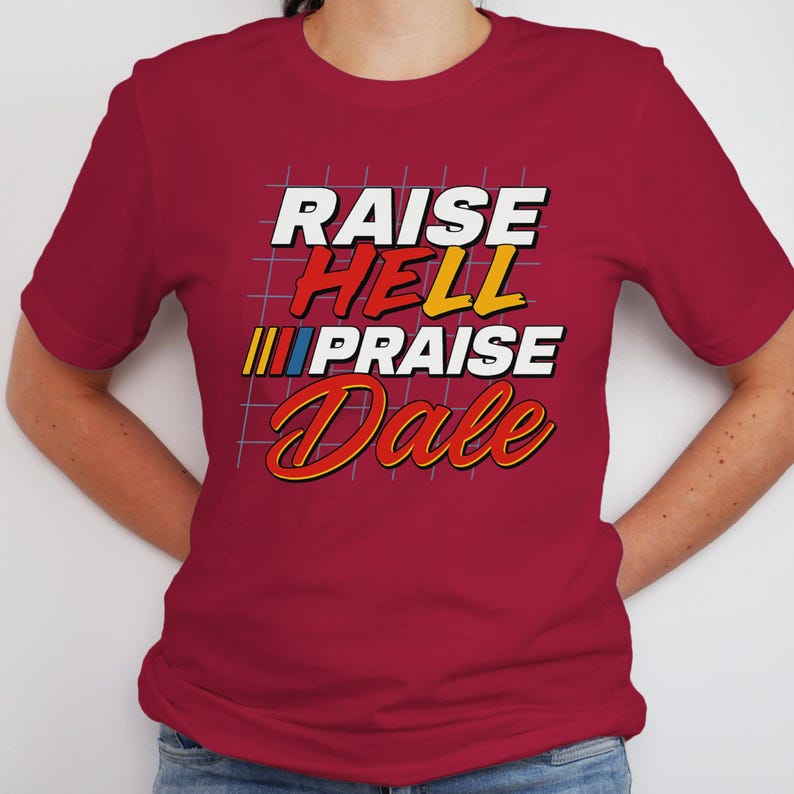 Pu&ograve; includere: Una maglietta bordeaux con la scritta "RAISE HELL PRAISE Dale" in un font retr&ograve;. Il testo &egrave; in bianco, rosso, giallo e blu. La maglietta ha maniche corte e girocollo.