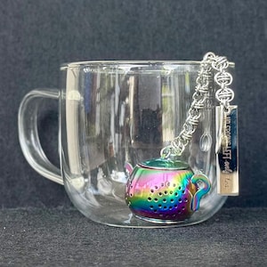 May include: A clear glass mug with a handle contains a rainbow-hued teapot-shaped tea infuser. The infuser is connected to a chain and a rectangular tag that reads "No crumbs left, only tea."