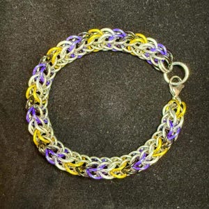 May include: A chainmail bracelet in silver, yellow, and purple. The bracelet is secured with a silver clasp and a large ring. The chainmail is made of interwoven metal rings. The bracelet's design is intricate and colourful.