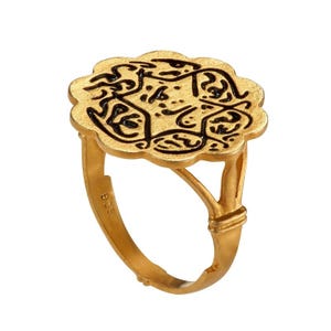 May include: A gold ring with a floral-shaped face. The face has black, stylized calligraphy. The ring's band is a simple, smooth design. The ring is set against a white background.