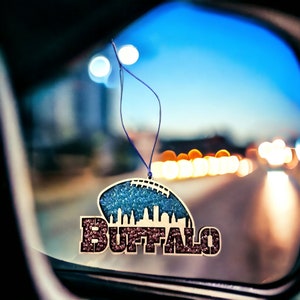 May include: A blue and brown car air freshener with the word "BUFFALO" in rhinestones. The air freshener is hanging from a rearview mirror in a car.
