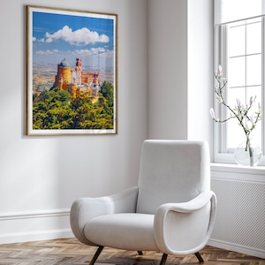 May include: A white armchair with a curved back and arms sits in a room with a framed picture of a yellow castle on a hill. The room has a white wall, a white baseboard, and a wood floor.