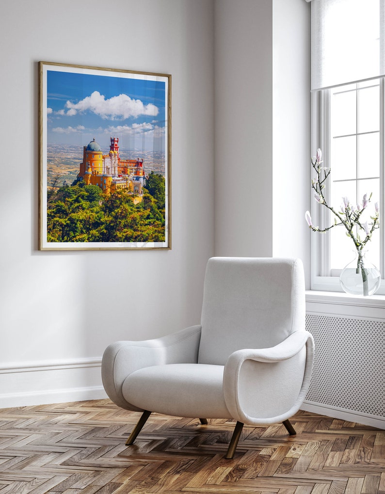 May include: A white armchair with a curved back and arms sits in a room with a framed picture of a yellow castle on a hill. The room has a white wall, a white baseboard, and a wood floor.