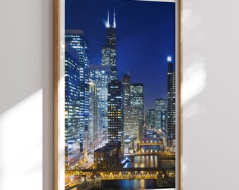 May include: A night-time aerial view of the Chicago skyline with tall buildings illuminated by lights. The Chicago River is visible in the foreground, with bridges crossing over it.