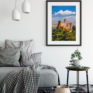 May include: A framed print of a yellow castle with a green roof and red accents, set against a blue sky with a white cloud. The castle is surrounded by green trees and is situated on a hilltop.