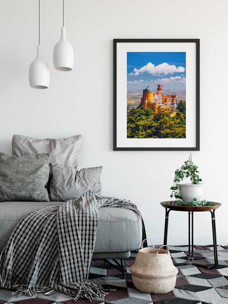 May include: A framed print of a yellow castle with a green roof and red accents, set against a blue sky with a white cloud. The castle is surrounded by green trees and is situated on a hilltop.
