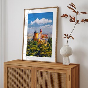 May include: A framed photograph of a yellow and red castle with a blue sky and white clouds in the background. The castle is surrounded by green trees.