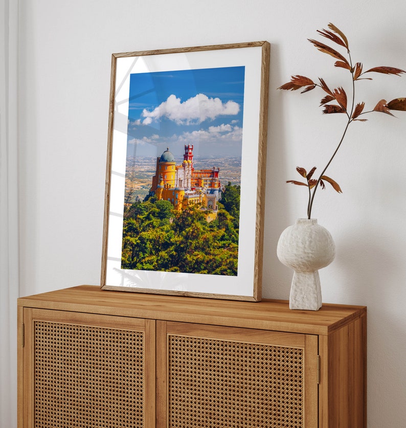 May include: A framed photograph of a yellow and red castle with a blue sky and white clouds in the background. The castle is surrounded by green trees.
