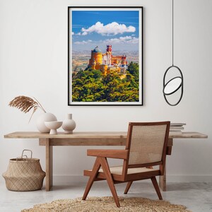 May include: A framed photograph of a yellow and red castle on a hilltop with a blue sky and white clouds in the background. The castle is surrounded by green trees.