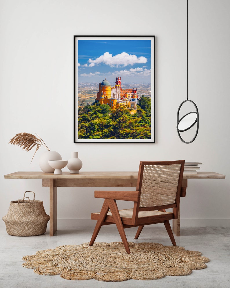 May include: A framed photograph of a yellow and red castle on a hilltop with a blue sky and white clouds in the background. The castle is surrounded by green trees.