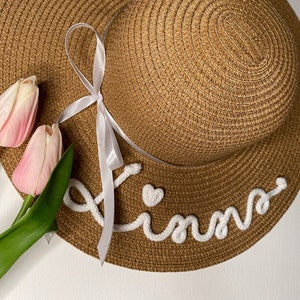 Può includere: Un cappello da sole di paglia marrone con tesa larga, un nastro bianco e il nome "Kinn" in corsivo bianco con un cuore. Due tulipani rosa con steli verdi sono sul lato sinistro del cappello. Un accessorio estivo.
