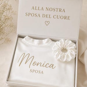Può includere: Una scatola regalo bianca contenente una camicia in raso bianco e un elastico per capelli bianco abbinato. L'interno del coperchio presenta il testo dorato ALLA NOSTRA SPOSA DEL CUORE con un cuore. La camicia riporta la scritta dorata Monica SPOSA.