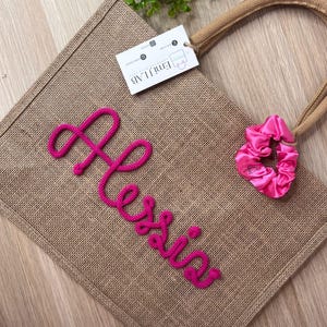 Può includere: Una borsa tote in tela di juta marrone con il nome "Alexis" in rosa ricamato sul davanti. Un elastico per capelli rosa e un'etichetta con il logo "Emi J Lab" sono attaccati. La borsa ha un manico in corda intrecciata.