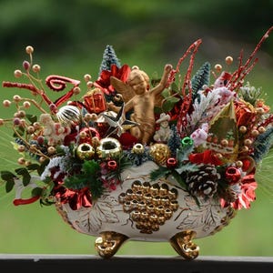 Peut inclure: Centre de table de Noël festif dans un bol décoratif blanc et doré. L'arrangement comprend un chérubin doré, des ornements rouges et dorés, des pommes de pin et des brins de verdure. Le bol repose sur trois pieds dorés.