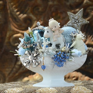 Puede incluir: Un cuenco blanco con pie rebosante de adornos navideños. El arreglo incluye adornos azules y plateados, una estrella y figuras de un niño a caballo y un oso polar. La base está decorada con cuentas azules y detalles plateados.