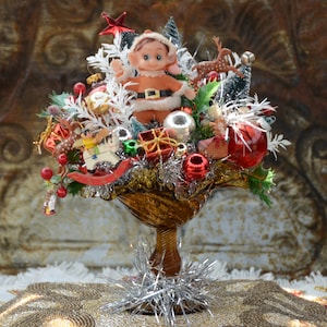 May include: A vintage Christmas centerpiece featuring a doll in a Santa suit, surrounded by ornaments, a red star, and a reindeer. The arrangement is in a gold-toned pedestal bowl, with silver tinsel accents. The background is a textured, ornate surface.