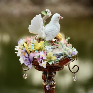 May include: A white porcelain dove figurine perched on a decorative stand adorned with colorful flowers. The stand is made of metal and has a vintage aesthetic.