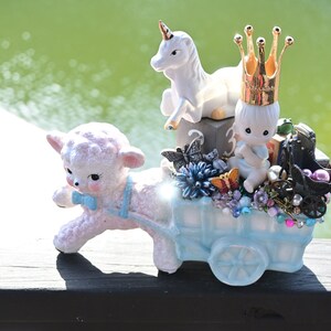 Op de afbeelding: Een wit keramiek lam trekt een blauwe wagen gevuld met verschillende decoratieve items, waaronder een wit eenhoorn, een babyfiguur met een gouden kroon en een zwarte koets met roze ballen. De wagen is versierd met vlinders, bloemen en juwelen.