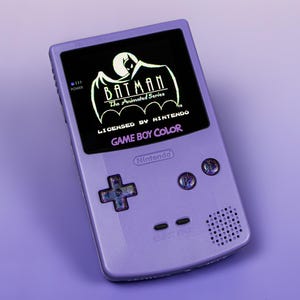 Könnte beinhalten: Ein lila Game Boy Color-Handheld-Konsolen-Gerät mit dem Logo von Batman: The Animated Series. Der Bildschirm zeigt das Batman-Logo mit dem Text "Batman The Animated Series" und "Licensed by Nintendo Game Boy Color".