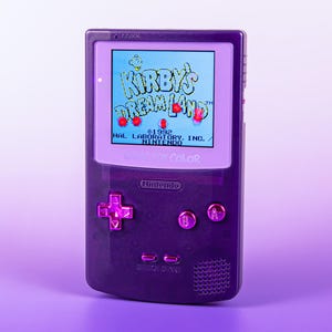 Könnte beinhalten: Ein lila Game Boy Color Handheld-Konsolen-Gerät, das das Spiel "Kirby's Dream Land" anzeigt. Der Bildschirm zeigt farbenfrohe Grafiken mit dem Titel des Spiels. Die Konsole hat ein Steuerkreuz, A- und B-Tasten sowie Start/Select-Tasten.