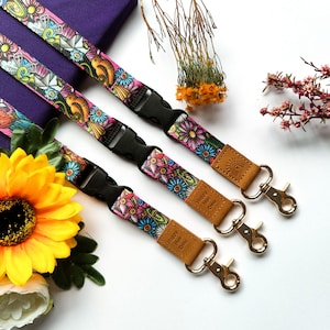 May include: Three colourful floral patterned lanyards with gold metal clasps. Each lanyard has a brown leather tag with the text "Happy Human Crew".