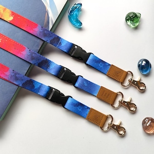 Puede incluir: Tres cordones coloridos con un patrón tie-dye azul, rojo y naranja. Cada cordón tiene una etiqueta de cuero marrón con el texto "Happy Place Crew" impreso en ella. Los cordones tienen clips y anillos de metal dorado.