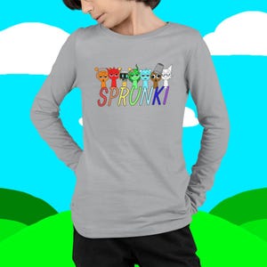 Puede incluir: Una camiseta gris de manga larga con la palabra "SPRUNKI" en colores arco&iacute;ris sobre una fila de personajes de dibujos animados. Los personajes son de varios colores y dise&ntilde;os. El fondo es un cielo azul y hierba verde.