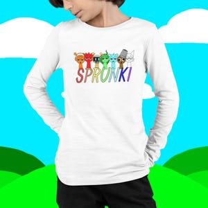 Puede incluir: Camiseta blanca de manga larga con la palabra "SPRUNKI" en letras coloridas y una fila de personajes de dibujos animados encima. Los personajes tienen varios colores y dise&ntilde;os, incluyendo naranja, rojo, verde y azul.